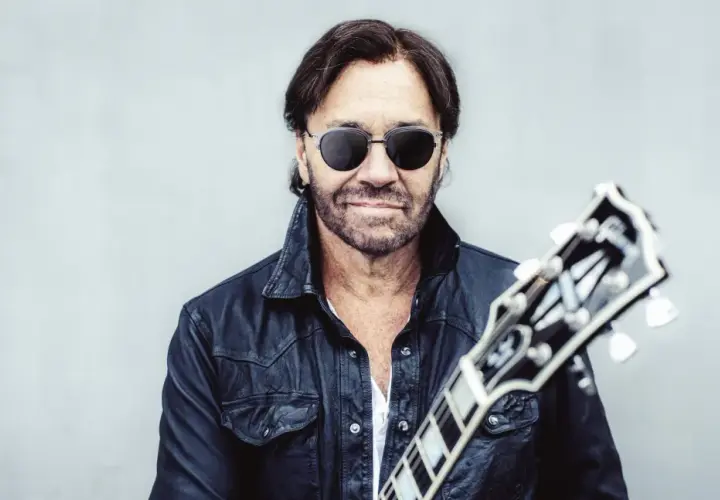 Al Di Meola w czarnej skórzanej kurtce i okularach przeciwsłonecznych, trzymający gitarę.