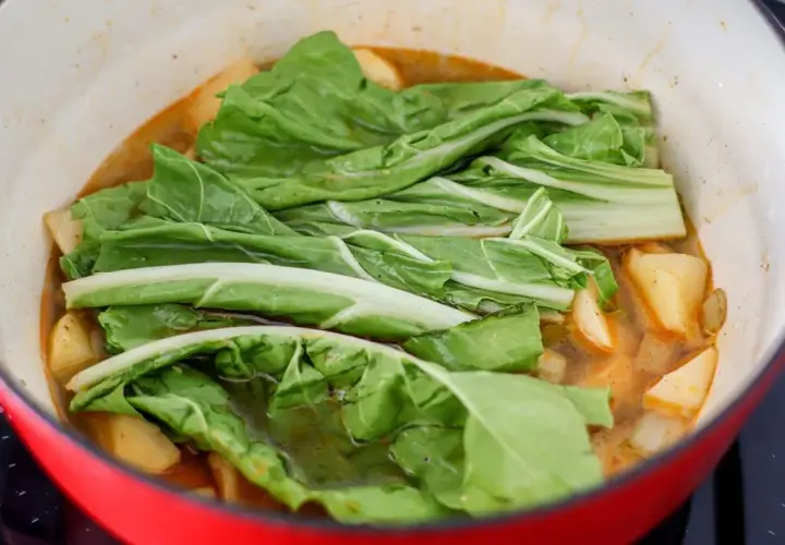 Verduras frescas, como acelgas, se cocinan en una olla roja con trozos de patata en un caldo sabroso.