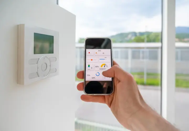 Control de la automatización de casas desde un smartphone. Se ve un termostato en la pared y una mano sujetando un teléfono con una app de control.