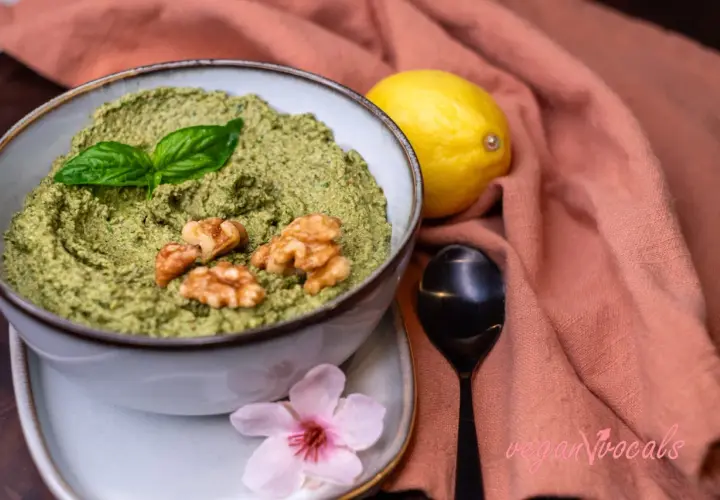 Un delicioso pesto vegano en un cuenco, adornado con hojas de albahaca y nueces. Junto a un limón y una flor rosa.