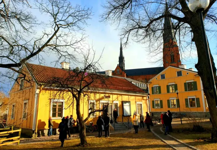 Gula hus med tegeltak och Uppsala domkyrka i bakgrunden. En grupp människor samlas utanför, kanske för ett besök på litteraturens hus Uppsala.