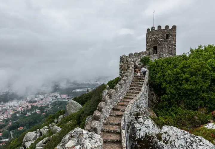 Escaleras de piedra ascienden hacia una torre en el **castillo de Sintra**, con vistas a la ciudad envuelta en niebla.