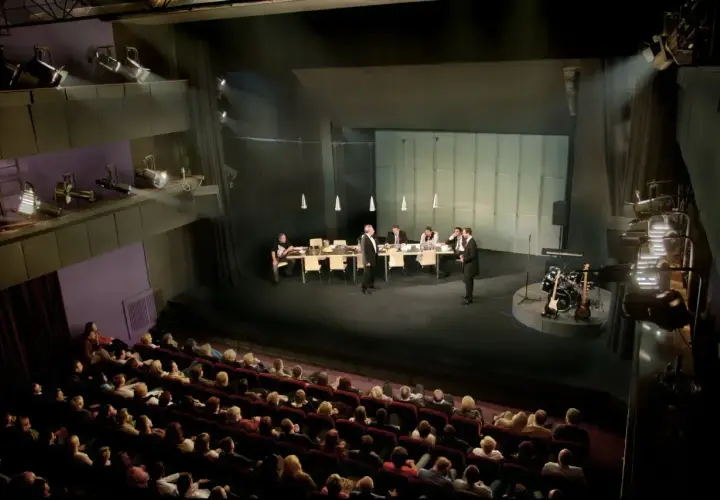 Scena teatralna z ludźmi przy stole i instrumentami muzycznymi. Widownia siedzi w ciemności, gotowa na spektakl. Dostawka w teatrze.