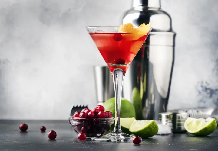 Elegancki Cosmopolitan przepis: czerwony drink w kieliszku, obok żurawina, limonka i shaker. Idealny na wieczór.