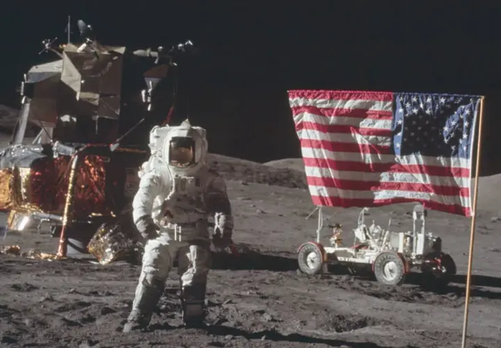 Astronaut w skafandrze podczas spaceru po księżycu, obok lądownika i łazika z amerykańską flagą.