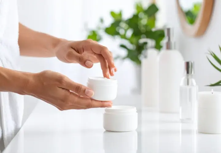 Dłonie nakładają krem z PEG-60 hydrogenated castor oil na twarz. W tle kosmetyki i rośliny.