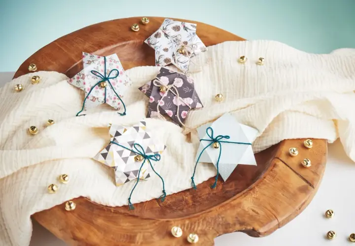 Fünf gefaltete Sternschachteln mit weihnachtlichen Mustern, verziert mit Schnur und Glöckchen, liegen auf einem cremefarbenen Tuch in einer Holzschale.