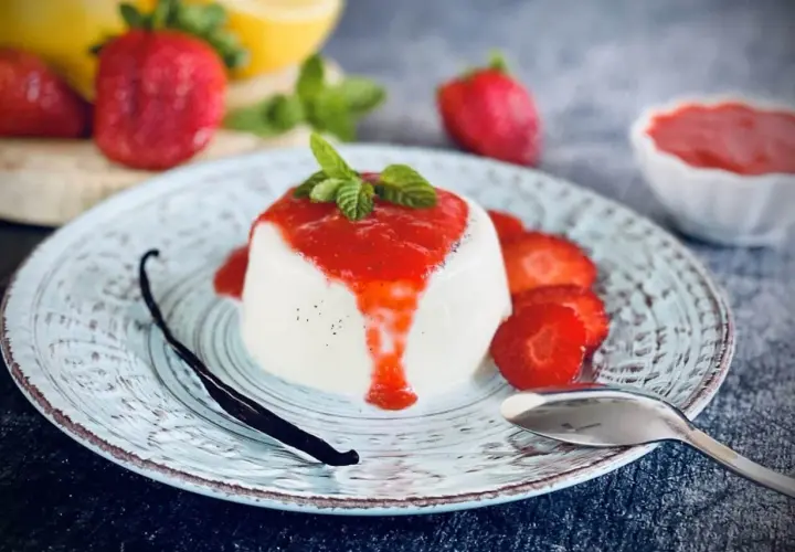 Cremige Panna Cotta mit Erdbeersauce, frischen Erdbeeren, Minze und einer Vanilleschote auf einem Teller.