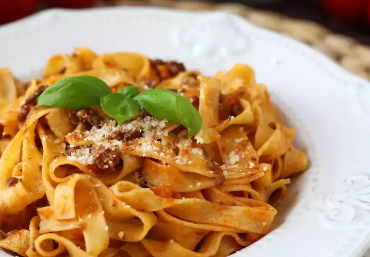 Pyszne tagliatelle bolognese z parmezanem i listkami bazylii na białym talerzu.