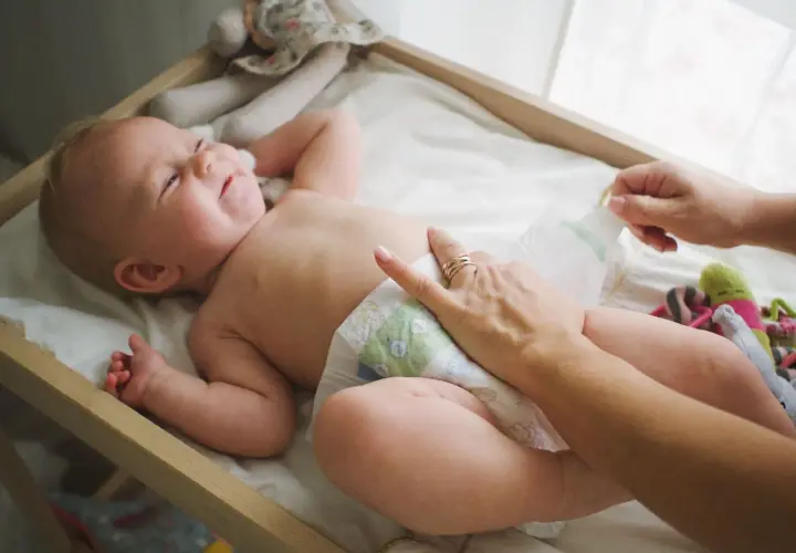 Radosna pielęgnacja niemowlaka chłopca. Mama zmienia pieluszkę, maluch uśmiecha się, leżąc na przewijaku wśród zabawek.