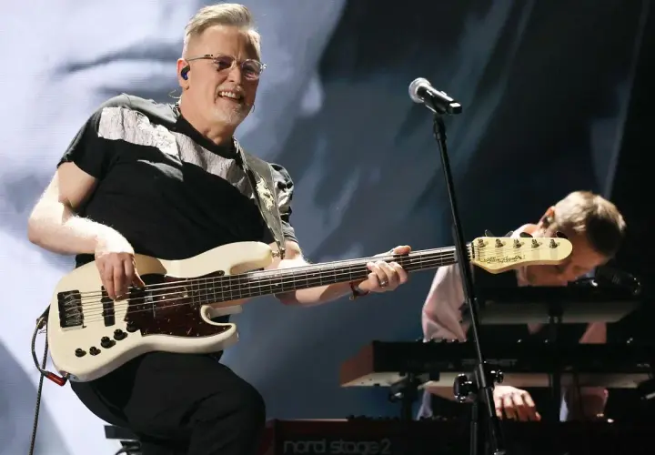 Muzyk gra na białej gitarze basowej, tworząc swoje opus magnum. W tle klawiszowiec pochyla się nad instrumentem.