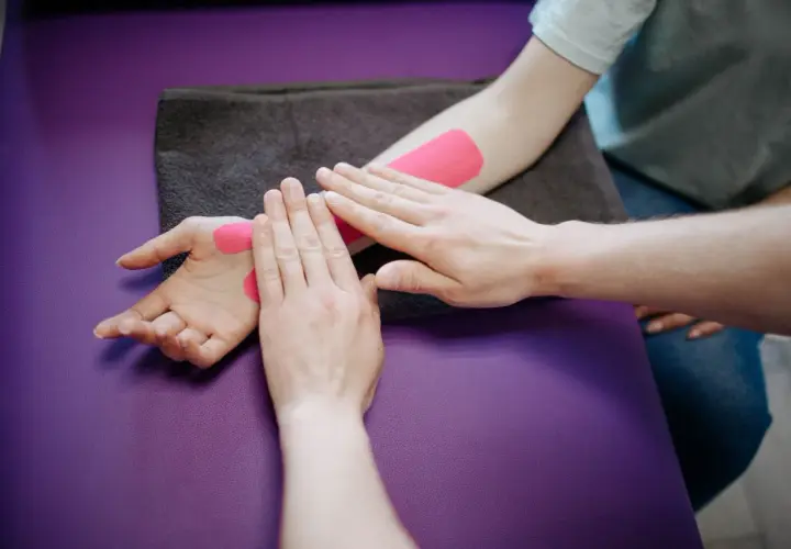 Fizjoterapeuta wykonuje slump test, aplikując różową taśmę kinezjotaping na przedramieniu i dłoni pacjenta.