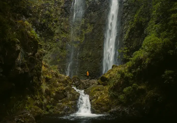 A lone hiker in an orange jacket stands at the base of the majestic Caldeirão Verde waterfall, a breathtaking sight on this lush hike.