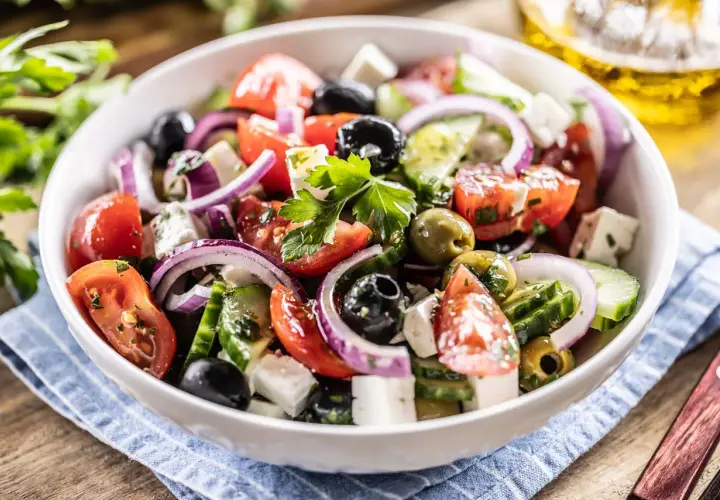 Ensalada griega fresca con tomates, pepinos, cebolla morada, aceitunas negras y queso feta, servida en un bol blanco.