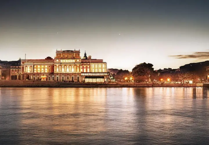 Kungliga operan i Stockholm lyser upp vid vattnet i skymningen, dess fasad speglas i det lugna vattnet.