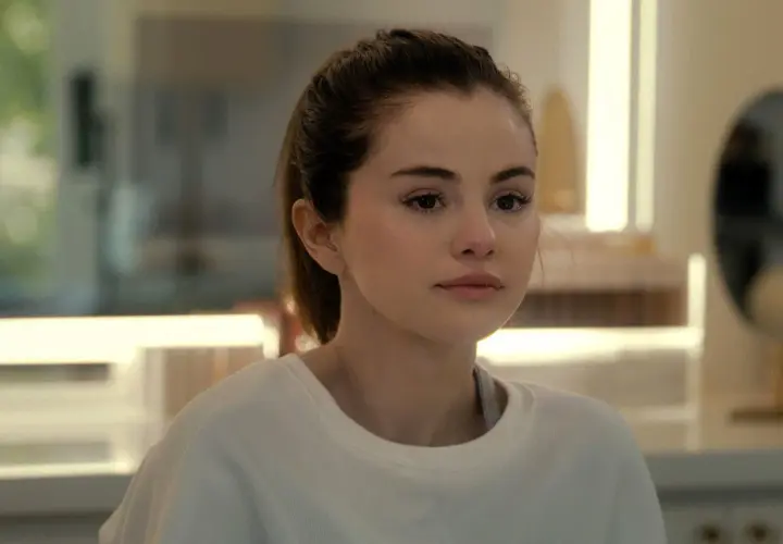 Selena Gomez w białej bluzie, z włosami związanymi w kucyk, patrzy w bok zamyślona.