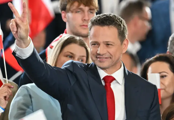 Rafał Trzaskowski w garniturze i czerwonym krawacie, pozdrawia tłum, unosząc rękę ze znakiem zwycięstwa.