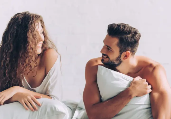 Coppia sorridente a letto, con la donna dai capelli ricci che guarda l'uomo con barba, un momento intimo prima di fare sesso.