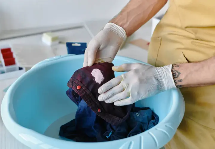 Hände mit Handschuhen behandeln ein Kleidungsstück mit einem rosa Fleck in einer blauen Schüssel, um **Flecken entfernen** zu können.