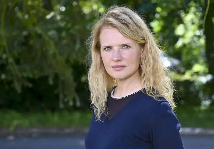 Wiktoria Grodecka z falowanymi blond włosami i niebieskimi oczami, w granatowym swetrze, na tle zieleni.