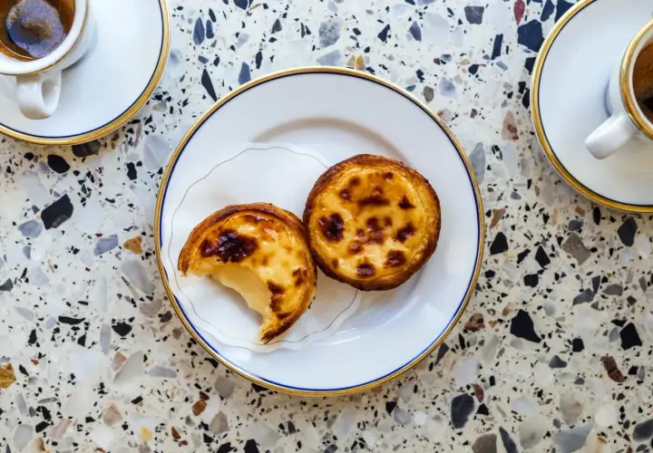 Dwa pyszne pastel de nata na talerzyku, obok filiżanki kawy. Idealny deser na popołudnie.