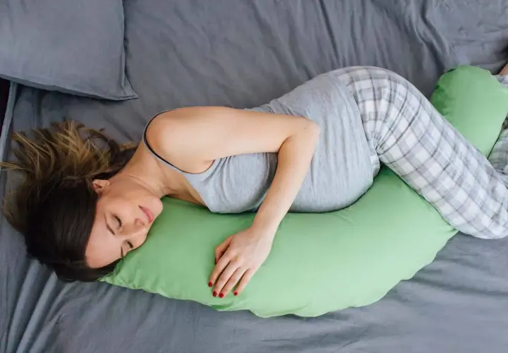 Donna incinta dorme su un fianco, supportata da un cuscino verde. Scopri come dormire in gravidanza per un riposo sereno.