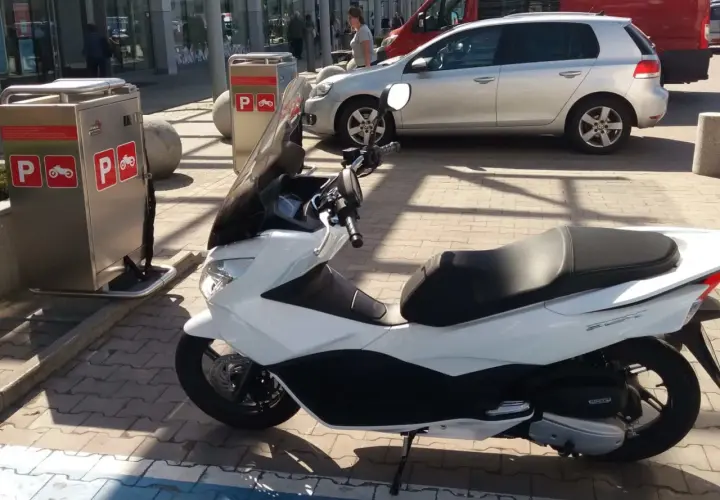 Na parkingu stoi biały skuter. Obok znajduje się automat z oznaczeniami "P" i symbolem motocykla, co sugeruje, że motocykle płacą za parking.