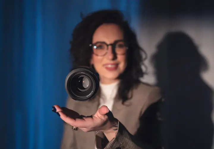 Uśmiechnięta kobieta w okularach unosi obiektyw aparatu nad dłonią. Czy fotograf przechowuje zdjęcia w obiektywie?