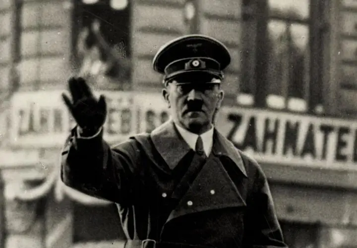 Adolf Hitler winkt. Er trug eine Uniform und einen Hut. Hinter ihm ist eine Tafel mit der Aufschrift "Zahnatelie". Was hat Hitler gemacht? Er hat den Zweiten Weltkrieg begonnen.