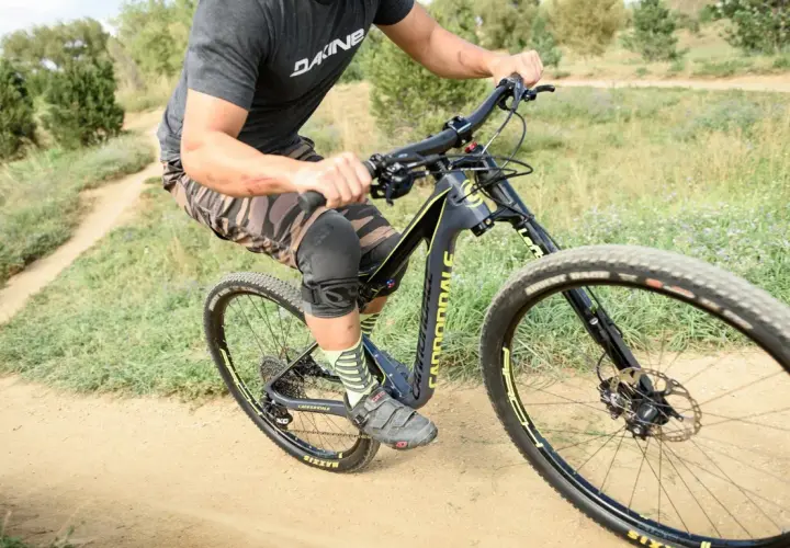 A rider on a black and yellow single fork mountain bike navigates a dusty trail, wearing knee pads and camouflage shorts.