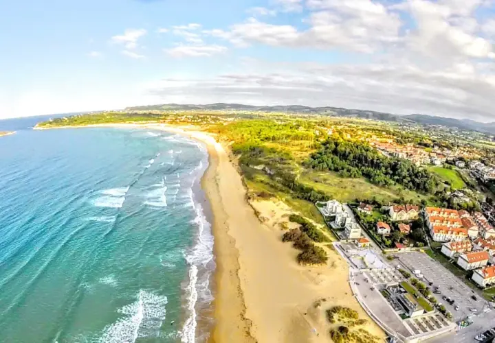 Vista aérea de la playa de Somo, con el mar turquesa, arena dorada, dunas y un pueblo costero con tejados rojos.