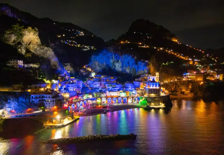 A vibrant night view of Vietri sul Mare, with buildings illuminated in colorful lights reflecting on the water.