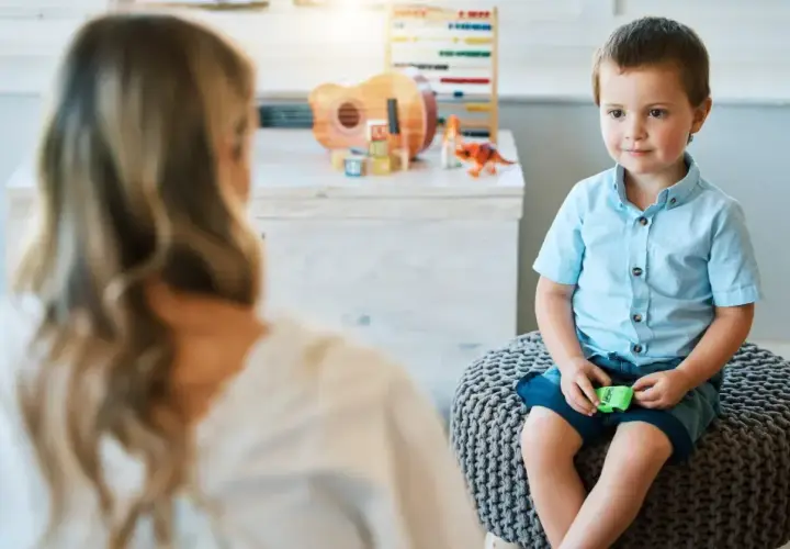Chłopiec w niebieskiej koszuli siedzi na pufie, rozmawiając z psychoterapeutką.