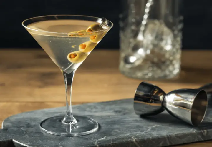 Un verre de martini gin, garni de trois olives, prêt à être dégusté. Un doseur à cocktail et un pichet à glaçons sont visibles en arrière-plan.