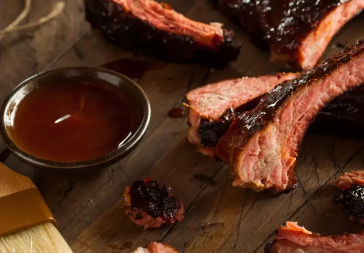 Soczyste żeberka BBQ, polane sosem, obok miseczka z marynatą do żeberek i pędzelek.