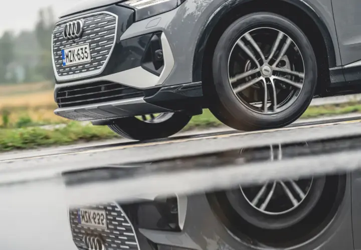 En Audi kör på en regnig väg, dess hjulbultar syns tydligt i den blöta asfalten som reflekterar bilen.