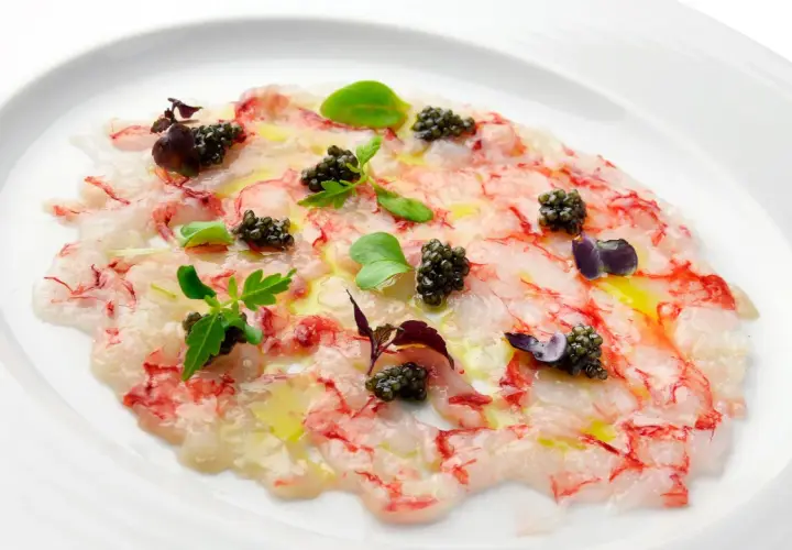 Delicado carpaccio de gambas con caviar, aceite de oliva y brotes verdes, presentado en un plato blanco.