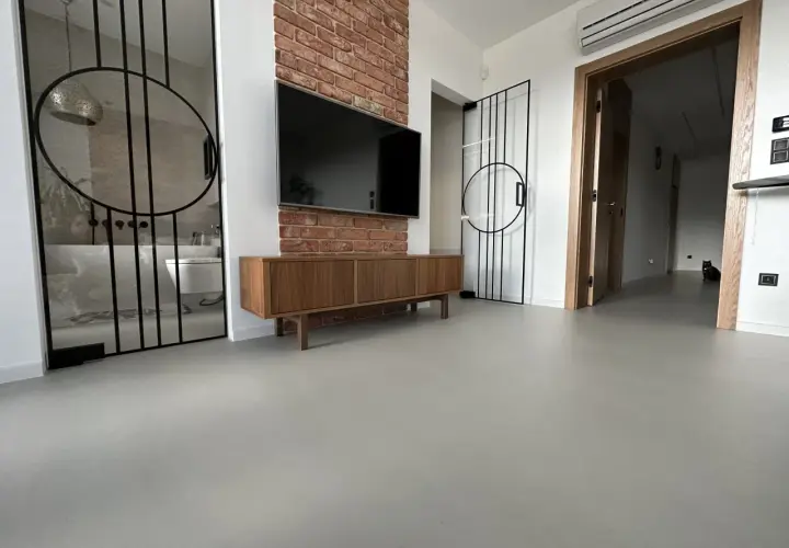 Nowoczesne wnętrze z betonową podłogą, co to jest mikrocement, która nadaje mu surowego charakteru. W tle widać łazienkę i salon.
