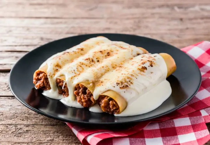 Tres canelones de carne gratinados con queso cremoso, servidos en un plato negro sobre un mantel a cuadros rojos.