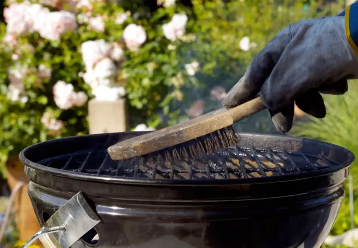 Main tenant gantée, brosse métallique pour nettoyer un barbecue. Des roses roses en arrière-plan.