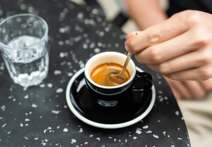 Ręka miesza łyżeczką aromatyczne espresso w czarnej filiżance. Obok szklanka wody. Prosty przepis na espresso jak zrobić.
