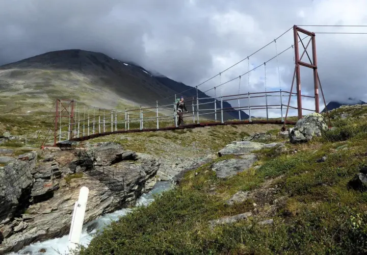 Hängebrucke über einem Fluss im schwedischen Gebirge. Ein Wanderer überquert die Brücke, umgeben von felsiger Landschaft und grüner Vegetation.