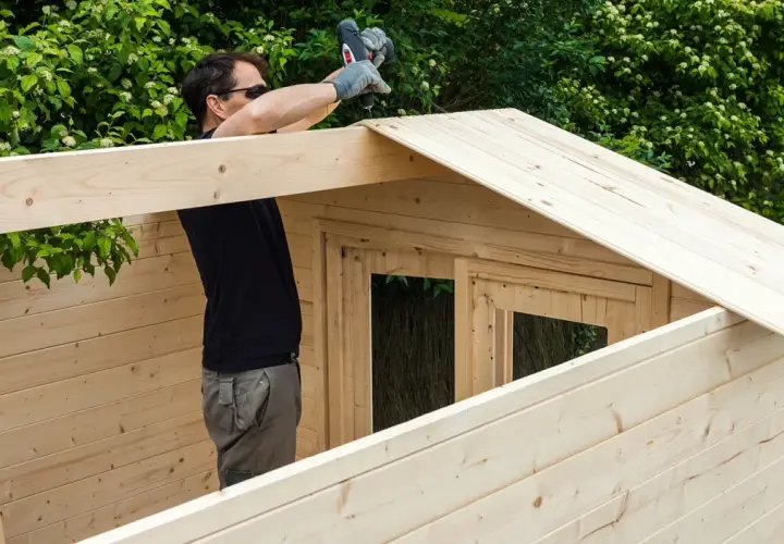 Un homme assemble une cabane en bois, utilisant une perceuse pour fixer une planche sur le toit. La peinture sur bois est visible sur les murs et le toit.