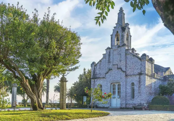 La iglesia de la Toja, cubierta de conchas, se alza majestuosa bajo un cielo azul con nubes. Un árbol frondoso y palmeras la rodean.