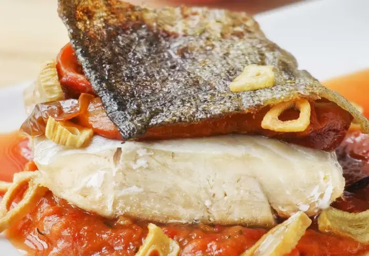 Bacalao a la riojana con piel crujiente, pimientos rojos y ajo frito, servido sobre una cama de salsa de tomate.