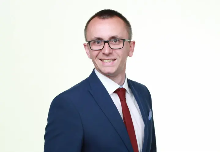Mężczyzna w okularach i granatowej marynarce z czerwonym krawatem, uśmiecha się.