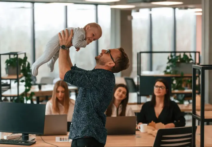 Szczęśliwy ojciec podrzuca dziecko w biurze, podczas gdy koleżanki pracują. Czy urlop wychowawczy wlicza się do stażu pracy?