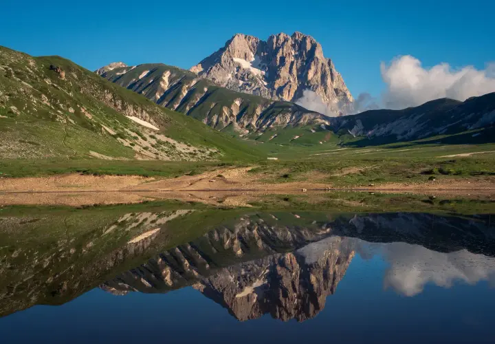 Spiegelglatte See reflektiert majestätisches **Gebirge in Italien** unter blauem Himmel. Grüne Hänge und felsige Gipfel.
