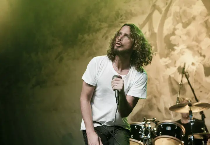 Chris Cornell z zespołem na scenie, w białej koszulce, śpiewa do mikrofonu. W tle perkusja i światła.