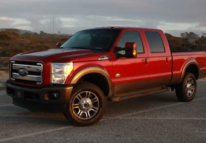 Czerwony Ford F350 King Ranch z brązowym pasem, zaparkowany na tle krajobrazu z wydmami i drewnianym płotem.